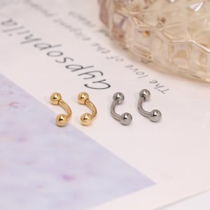 May include: Four curved barbell earrings. Two are gold-colored, and two are silver-colored. Each earring has a small ball on each end. The earrings are displayed on a white surface with text in the background.