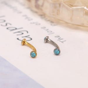Könnte beinhalten: Zwei gebogene Augenbrauenpiercings. Eines ist goldfarben, das andere silberfarben. Jeder Ring hat einen kleinen, runden, türkisfarbenen Stein am Ende. Der Schmuck wird auf einer weißen Oberfläche mit schwachem Text im Hintergrund präsentiert.