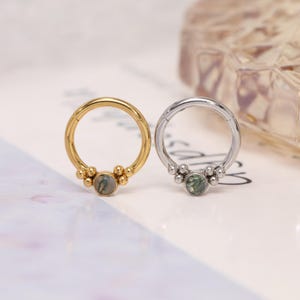 May include: Two circular hinged hoop rings, one gold-toned and one silver-toned. Each ring features a central round moss agate gemstone flanked by a cluster of three small metallic spheres on both sides, creating a symmetrical decorative design.