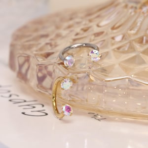 May include: Two open-ended hoop earrings, one in a silver tone and one in a gold tone. Each earring features two round, iridescent, multi-colored stones set at the open ends of the circular metal band, creating a reflective and shimmering effect.