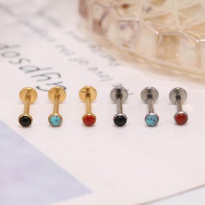 Puede incluir: Seis piercings labiales con parte trasera plana. Tres son de color dorado y tres plateados. Cada uno tiene una pequeña gema redonda en negro, turquesa o rojo. Los piercings se muestran sobre una superficie blanca.