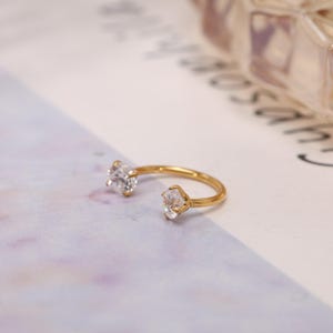 May include: A gold-colored ring with two clear, sparkling gemstones. The ring has an open design, with the gemstones set at opposite ends of the band. The ring is displayed on a soft, patterned surface.
