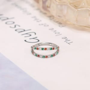 May include: A silver double-band ring with alternating red, green, and clear gemstones. The ring is set on a white surface with the word "Gypsop" and other text visible in the background.