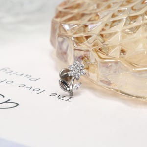 May include: A silver flower body jewelry with clear cubic zirconia stones. The flower has a stem and leaf.