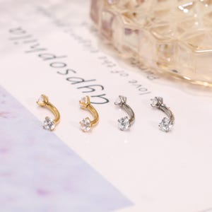 May include: Four curved eyebrow rings. Two are gold-toned, and two are silver-toned. Each ring features a clear, round gemstone at each end. The jewelry is displayed on a white surface with blurred text in the background.