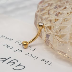 May include: Gold body jewelry with a clear gem on each end. The jewelry is curved and is likely a belly button ring.