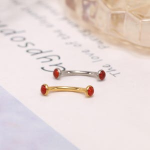 Könnte beinhalten: Zwei gebogene Barbells, eine goldfarbene und eine silberfarbene, jeweils mit einem kleinen, runden, roten Edelstein an jedem Ende. Der Schmuck wird auf einer weißen Oberfläche mit schwachem Text im Hintergrund präsentiert.