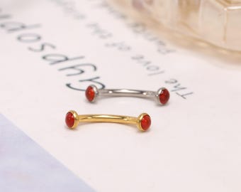 Könnte beinhalten: Zwei gebogene Barbells, eine goldfarbene und eine silberfarbene, jeweils mit einem kleinen, runden, roten Edelstein an jedem Ende. Der Schmuck wird auf einer weißen Oberfläche mit schwachem Text im Hintergrund präsentiert.