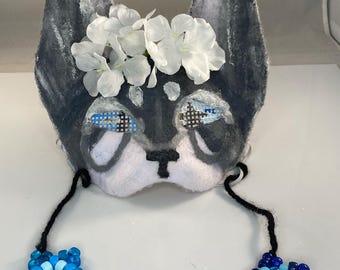 May include: A handmade, gray and white felt mask in the shape of a dog's face, adorned with white floral accents. The mask features blue beaded accents around the eyes and dangling star-shaped beaded embellishments. The mask is designed for costume or decorative use.