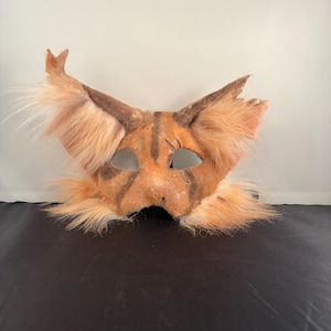 May include: A handmade cat mask with orange and brown fur, featuring pointed ears and dark brown stripes. The mask has eye cutouts and is designed to be worn. The fur is long and fluffy, giving it a realistic appearance.