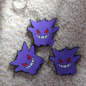 Puede incluir: Tres llaveros de Gengar morados con ojos rojos y dientes blancos. Un llavero está unido a un llavero plateado. Los llaveros están sobre una superficie texturizada blanquecina. Un accesorio divertido.