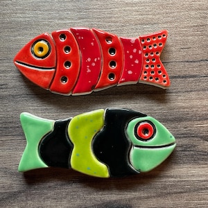 May include: Two ceramic fish ornaments. One is red with a yellow eye and black dots. The other is green, black, and yellow with a red eye. Both have a glossy finish and are arranged on a wooden surface.