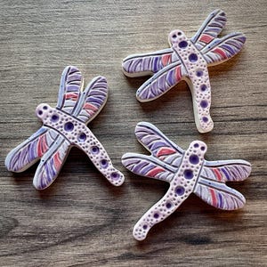 May include: Three ceramic dragonfly figurines. Each dragonfly has a white body with purple, red, and blue wings. The bodies are dotted with purple circles. The figurines are arranged on a wooden surface.