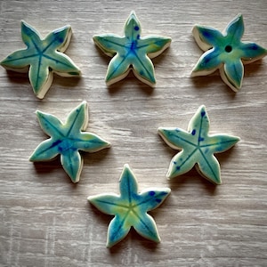 Peut inclure: Six étoiles de mer en céramique avec un glaçage turquoise et bleu. Chaque étoile de mer a un design à cinq pointes avec un bord blanc. Les étoiles de mer sont disposées sur une surface à grain de bois clair.