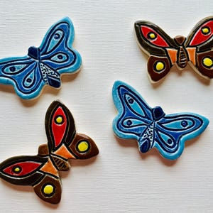 May include: Four ceramic butterfly decorations in shades of blue, brown, red, orange, and yellow. The butterflies have detailed wing patterns and are arranged on a white surface. These decorative butterflies are suitable for home decor.