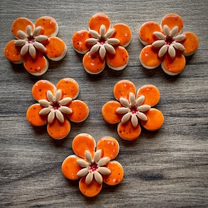 May include: Six ceramic flower-shaped tiles. Each tile has five orange petals with dark speckles, a white edge, and a beige centre with a red accent. The tiles are arranged on a wooden surface.
