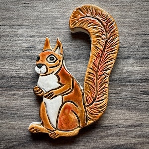 May include: A white ceramic mug with a black handle and the text "But First, Coffee" in a bold, black font. The mug is set against a blurred background. The mug appears to be approximately 10 cm tall.