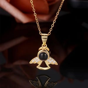 May include: Gold-colored angel pendant necklace with a black center stone and small clear stones on the wings. The chain is also gold-colored and has a delicate link design. The necklace is displayed against a dark background.