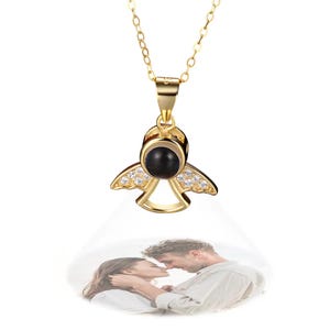 May include: A gold-colored angel pendant necklace with a black center stone and clear accent stones on the wings. The pendant is suspended from a delicate chain. A projected image of a couple is visible below the pendant.