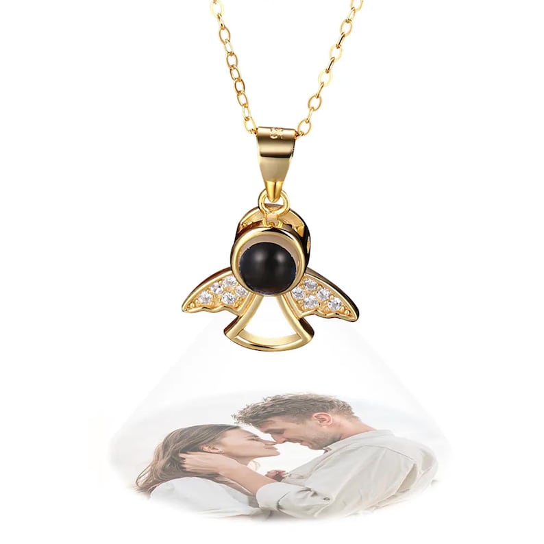May include: A gold-colored angel pendant necklace with a black center stone and clear accent stones on the wings. The pendant is suspended from a delicate chain. A projected image of a couple is visible below the pendant.
