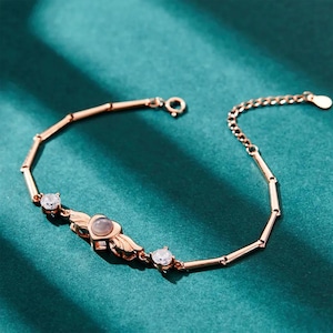 Op de afbeelding: Roségouden armband met een hartvormige bedel en twee ronde, heldere edelstenen. De armband heeft een delicaat kettingontwerp en wordt gepresenteerd op een teal groene ondergrond. De hartbedel heeft kleine vleugels aan weerszijden.