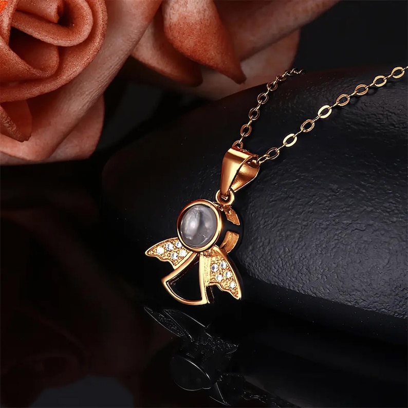 May include: A gold-colored angel pendant necklace. The pendant features an angel design with a central, circular stone and wings adorned with small, clear crystals. The necklace chain is delicate and gold-toned.