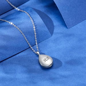May include: A silver teardrop-shaped pendant necklace with a small opening on the side. The pendant is engraved with the initials "R.I.P."