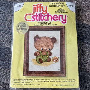 May include: A Jiffy Stitchery kit, titled "Cuddly Cub," features a cross-stitch design of a teddy bear wearing green dungarees. The kit includes a printed design, yarn, needle, and instructions. The packaging is yellow with the price £2.39 in the upper left corner.