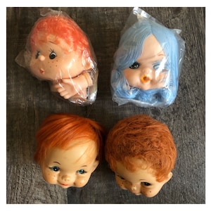 May include: Four vintage doll heads with red, orange, and blue hair. Two are in plastic packaging. The doll heads have painted faces, including freckles, and are arranged on a wooden surface.