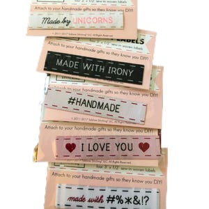 May include: A stack of pink and white woven labels with various phrases in black and pink text. Phrases include "Made by UNICORNS", "MADE WITH IRONY", "#HANDMADE", "I LOVE YOU", and "made with #%*&!?". Each label is approximately 3 x 1/2 inches.