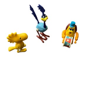 May include: Three cartoon character figurines. A yellow bird with black eyes and a small beak. A blue and white roadrunner with a yellow beak and dark blue crest. A yellow dog with a blue hat and a red, white, and blue sign.
