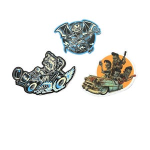 Op de afbeelding: Drie stickers met illustraties. Eén toont een skelet dat gitaar speelt met vleermuisvleugels, een ander een skelet dat een hot rod bestuurt, en de laatste een band in een oldtimer.