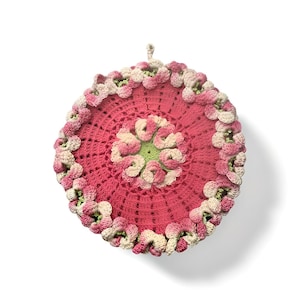 Può includere: Un sottopentola rotondo fatto a mano, all'uncinetto, nei toni del rosa, crema e verde. Il centro presenta un motivo floreale, circondato da un bordo di piccoli fiori tridimensionali. Un anello è attaccato per appendere.