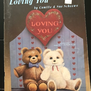 May include: A book cover titled "Loving You" by Camille & Sue Scheewe. The cover features a heart-shaped design with the words "Loving You" and illustrations of two teddy bears. The background is a light blue with red hearts.