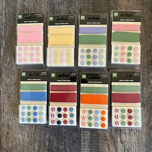 May include: Eight packs of ribbon and ribbon brads. Each pack contains three rolls of ribbon with different patterns and 12 matching brads. The ribbons are in various colours and patterns, including pink, yellow, green, blue, red, orange, and brown. The brads are in matching colours and patterns.