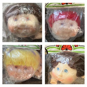 Op de afbeelding: Vier vintage poppenhoofden met verschillende haarkleuren: bruin, rood, geel en donkerbruin. Elk poppenhoofd is verpakt in een doorzichtige plastic zak. De poppenhoofden hebben geschilderde gezichtskenmerken, waaronder blauwe ogen en roze wangen.