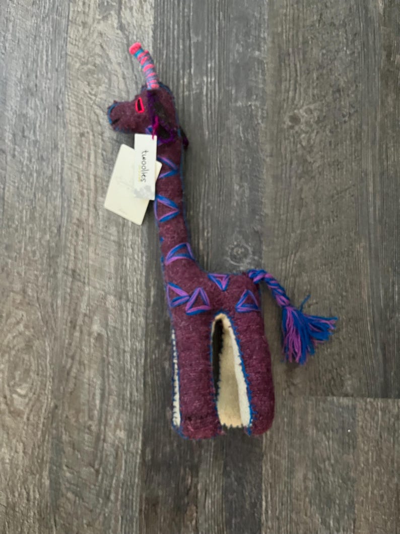 Pode incluir: Um brinquedo de girafa de pel&uacute;cia feito &agrave; m&atilde;o em tons de roxo, azul e rosa. A girafa tem um pesco&ccedil;o longo e uma cauda tran&ccedil;ada. A etiqueta diz "Twoolies".