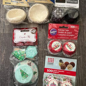 May include: Assortment of baking cups and cupcake kits in various designs. Includes white, black, and patterned paper cups, along with a cupcake kit featuring picks. The image showcases a variety of sizes and decorative styles for baking.