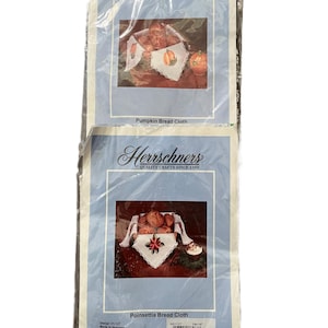 May include: Two Herrschners bread cloth kits in clear plastic packaging. The top kit features a pumpkin design, and the bottom kit features a poinsettia design. Each package displays a finished cloth with lace edges and includes the text Pumpkin Bread Cloth and Poinsettia Bread Cloth.