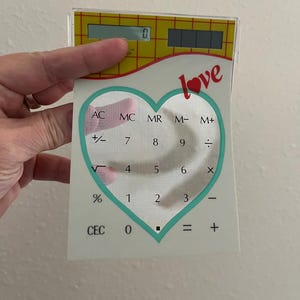 Può includere: Una calcolatrice trasparente a forma di cuore con un contorno turchese e la parola "love" in rosso. La calcolatrice ha una parte superiore gialla con un display digitale e pulsanti numerici.