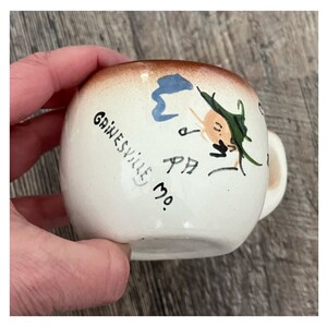 May include: A small, white ceramic mug with a brown rim and a small handle. The mug is decorated with a whimsical face and the text "Gainesville PA MO."