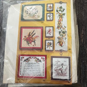 Op de afbeelding: Een pakket met verschillende kruissteekpatronen. De patronen tonen bloemstukken, vogels en een lange banner met een bloemmotief. Eén patroon bevat de tekst "God grant me the serenity..." in een decoratief kader.