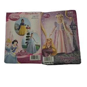 Può includere: Due buste di modelli di cucito Simplicity con principesse Disney. Un modello è etichettato "Simplicity 2817" e l'altro è "Simplicity 2065" con la parola "Tangled". I modelli mostrano illustrazioni di abiti da principessa.