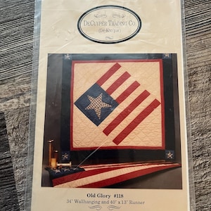 以下が含まれることがあります： 愛国的なテーマのキルトパターンが特徴のパッケージ。デザインには、クリーム色の背景に青い星と赤いストライプが含まれています。「Old Glory #118」と書かれており、86 cmの壁掛けと102 cmのランナーを示しています。