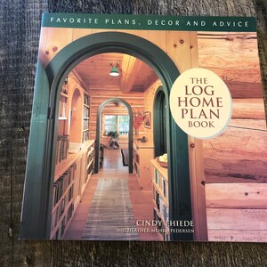 Puede incluir: El libro de planos de casas de madera de Cindy Thiede, con una fotografía del interior de una casa de madera. La portada incluye el texto « FAVORITE PLANS, DECOR AND ADVICE ». El diseño del libro muestra un interior acogedor y acogedor de cabaña de madera con arcos y estanterías.