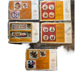 May include: A selection of vintage cross-stitch kits with diverse designs. The kits showcase images of mushrooms, Christmas ornaments, a house with a car, and a panda bear. Each kit is packaged in a rectangular box with the brand name "The Creative Circle".