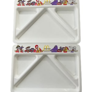 May include: Two white plastic divided plates featuring a colorful border with McDonald's characters. Each plate has a large compartment and two smaller sections, ideal for separating food items. The McDonald's logo is also visible.