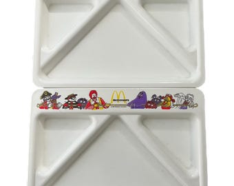Może przedstawiać: Dwa białe plastikowe talerze z przegródkami z kolorową obwódką z postaciami z McDonald's. Każdy talerz ma dużą komorę i dwie mniejsze sekcje. Widoczne jest również logo McDonald's.