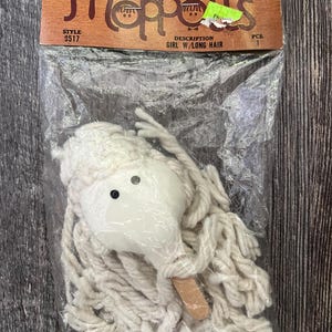 May include: A white cotton mop head doll with black eyes and a wooden stick. The doll is packaged in a clear plastic bag with a label that reads "Westrim Crafts Mini Moppets Style 9517 Description Girl w/Long Hair".