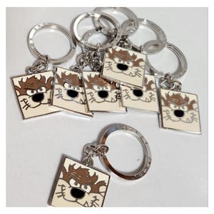 Op de afbeelding: Een verzameling zilveren sleutelhangers met een vierkante bedel met een cartoonkarakterontwerp. De bedel heeft een witte achtergrond met een bruin en zwart Tazmanian Devil-gezicht. De sleutelhangers hebben een zilveren ring.